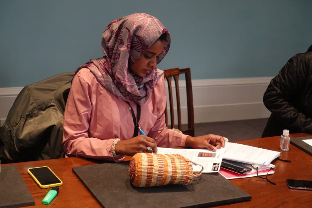 A woman in pink clothes and head scarf sits examining a museum object