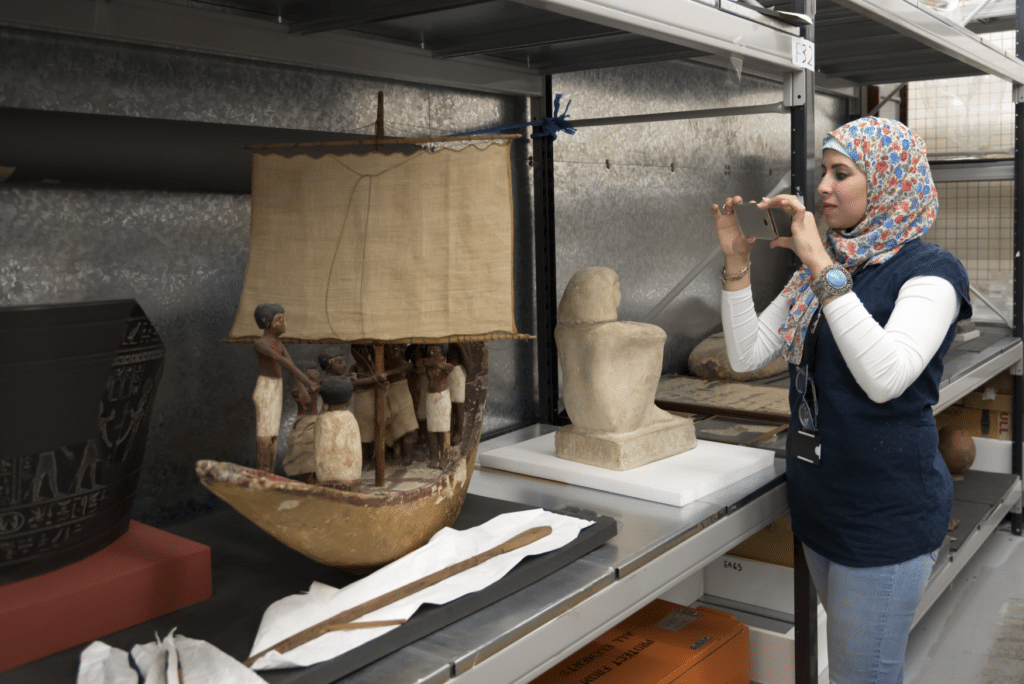 Heba Khairy in stands in a storage room at the British Museum and is taking a photograph of an Egyptian object shaped like a boat.