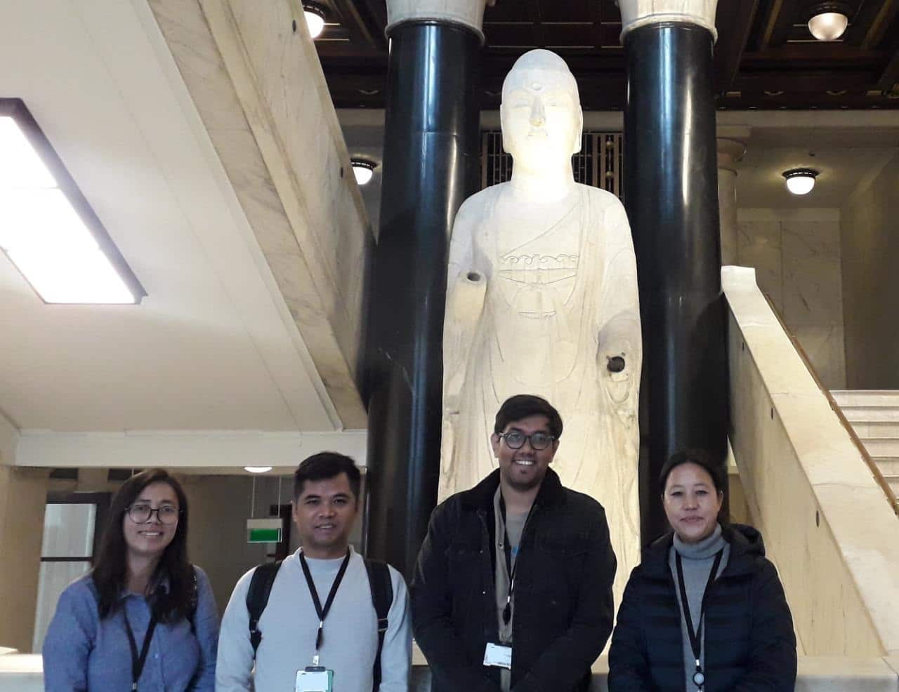 Four people stand in a line in front of a large statue depicting a human figure