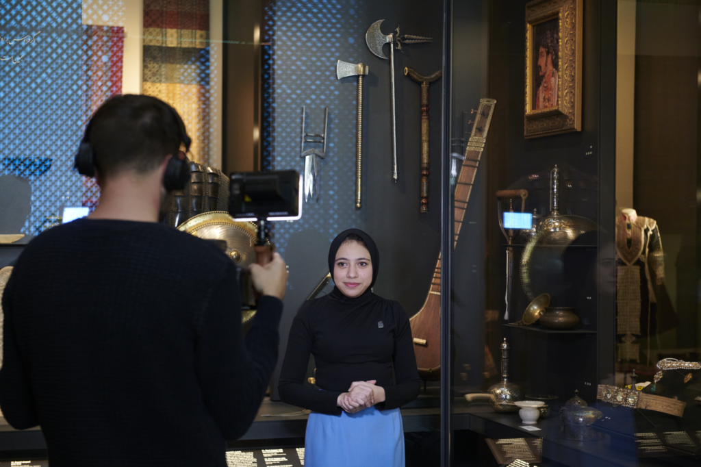 Dana Khalil stands in front of a display case preparing to film her presentation. The cameraman faces away from the photo holding a lights towards Dana.