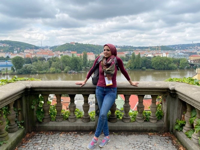 Noor standing on a terrace with a view of Prague in the background.