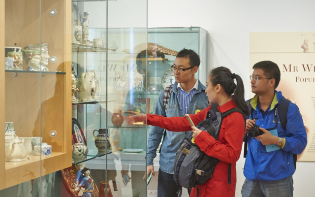 Three people looking at a museum gallery case, one person is pointing at an object in a case.
