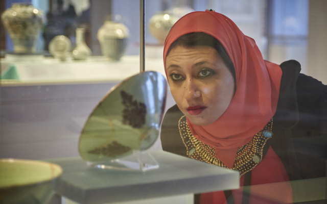 A woman wearing a pink headscarf looks through a glass display case at a ceramic bowl inside.