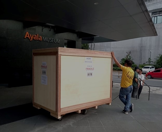 Photograph of a large crate being wheeled by a man outside the Ayala Museum.