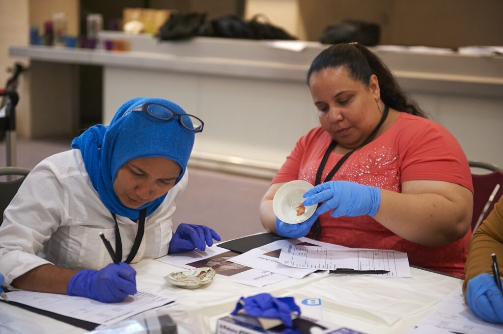 Mariem sat at a table holding a ceramic bowl whilst wearing blue gloves. Another woman sits beside her, writing.