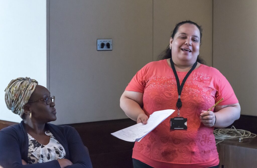 Mariem (right) stood holding a piece of paper and presenting. A woman sits beside her to the left.