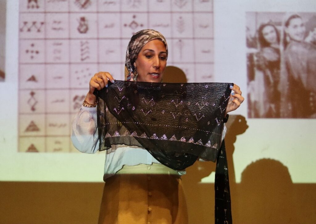 Photograph of a woman giving a presentation. She is holding a piece of black fabric embellished with beads.