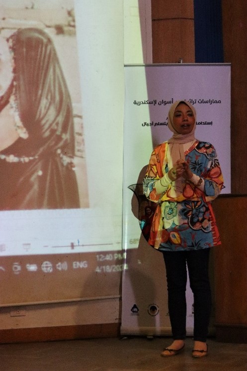 Photograph of a woman standing in front of a projector. She is speaking to an audience who are not in shot.