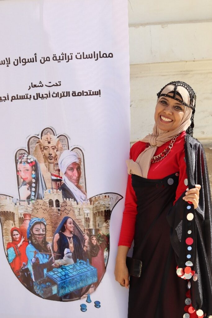 A photograph of a woman wearing traditional regional Egyptian dress smiling in front of a poster.