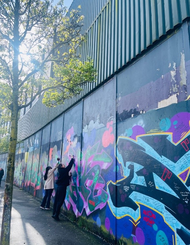 Photograph of two women standing against the Belfast peace wall, writing a message.