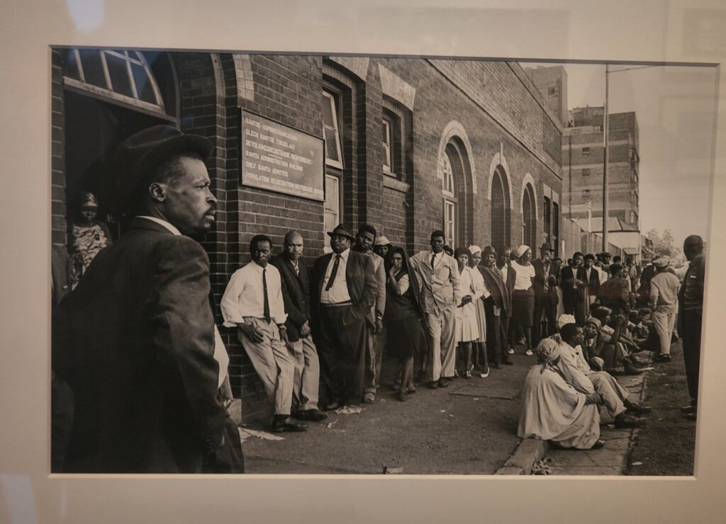 Photograph of a work by Ernest Cole of black people queuing outside a train station
