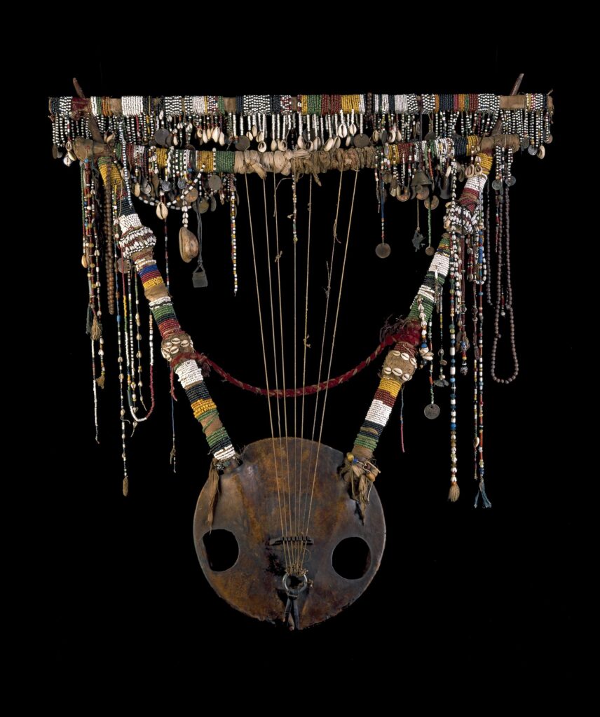 Sudanese Lyre. Large musical instrument, lyre, with two 'arms' set into a semi-spherical resonator of wood covered with skin, into which two circular apertures have been cut, giving the appearance of a face. Seven strings are attached to the resonator and are wound around the first of two cross pieces, both of which are covered with an assortment of pendant charms, coins and beads.