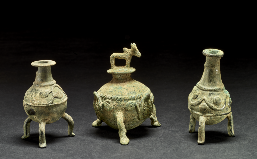 Copper-alloy cosmetic container; cast; spherical pot on three bull's legs, three leaves held up by scrolling between legs; coiled cable around vessel body; neck and mouth at top of vessel, stopper with lid in the form of a standing humped bovine.