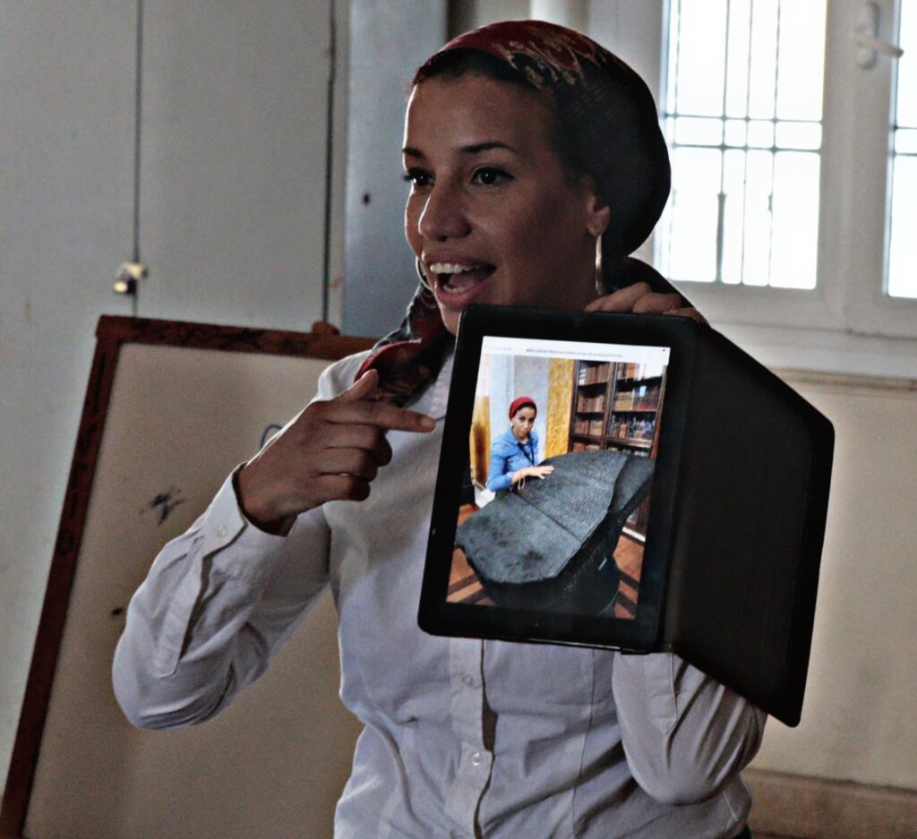 A woman holds up an iPad with a photograph of her touching a replica Rosetta Stone in the British Musuem.