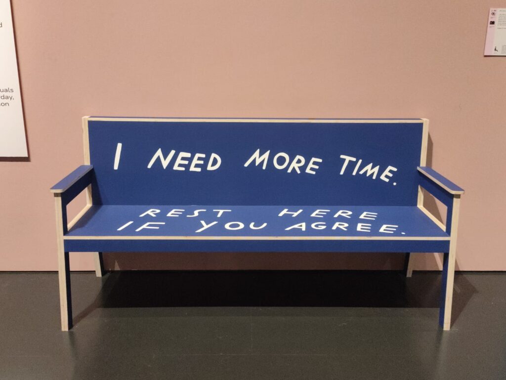 Photograph of a blue bench which reads: 'I need more time. Sit here if you agree.