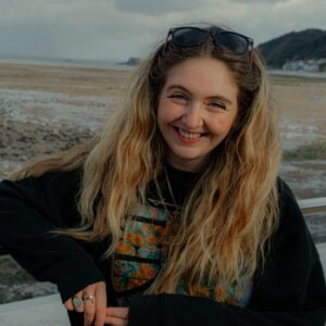 Photograph of Caitlin, wearing a black jumper, with the sea in the background.