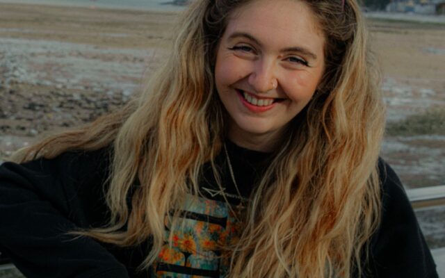 Photograph of Caitlin, wearing a black jumper, with the sea in the background.
