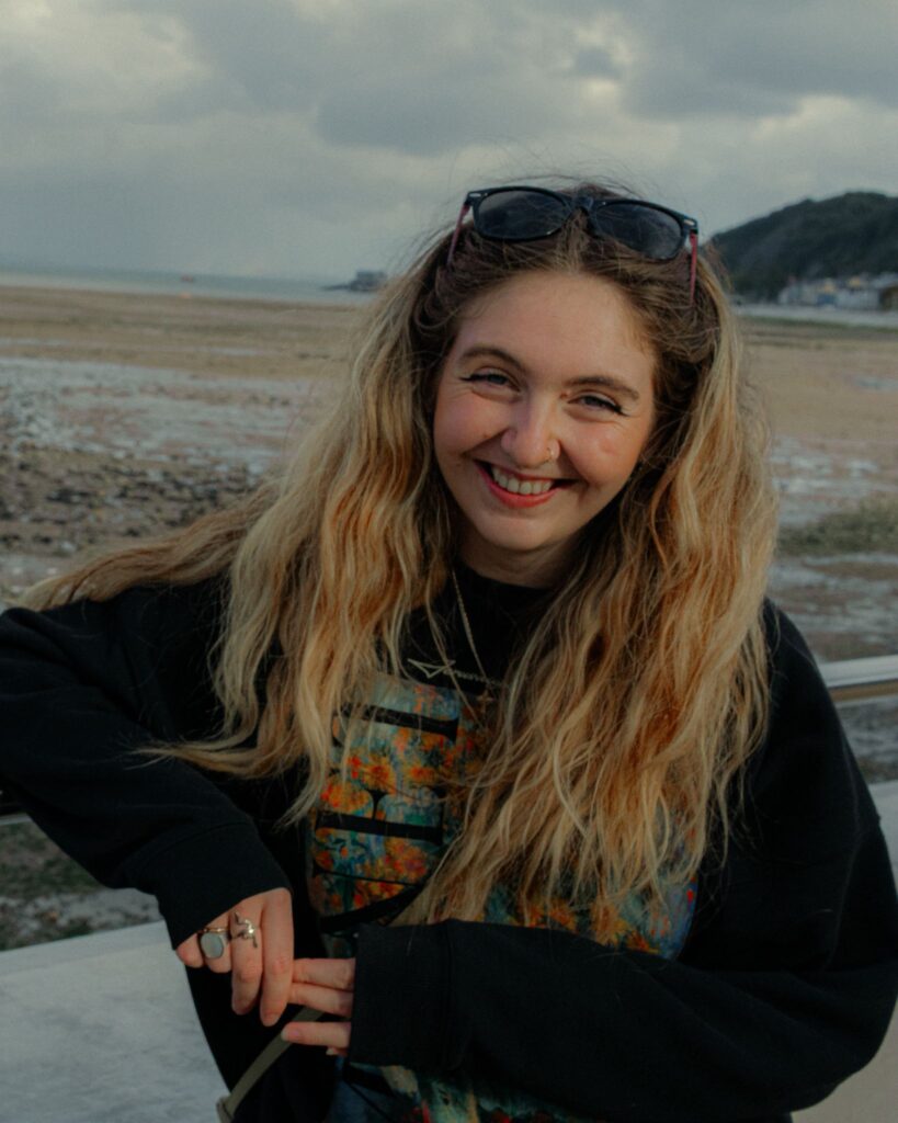 Photograph of Caitlin, wearing a black jumper, with the sea in the background.